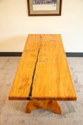 A rectangular wooden table with a live edge and a prominent black resin crack running down the center, supported by a chunky carved wooden base, set on a carpeted floor against a beige wall.