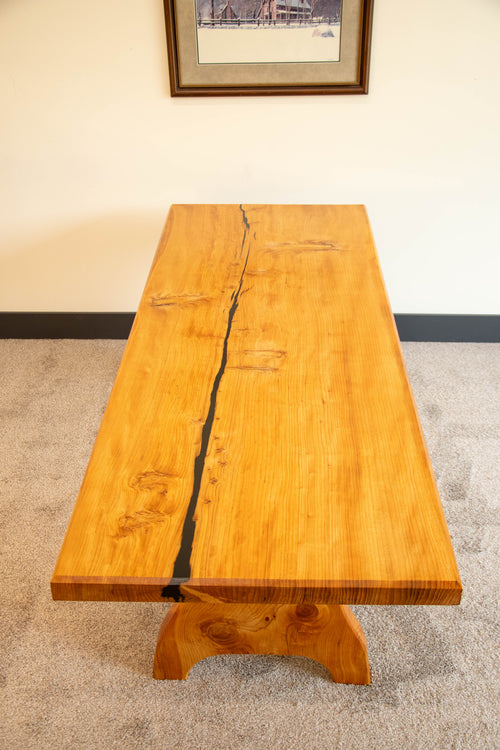 A rectangular wooden table with a live edge and a prominent black resin crack running down the center, supported by a chunky carved wooden base, set on a carpeted floor against a beige wall.